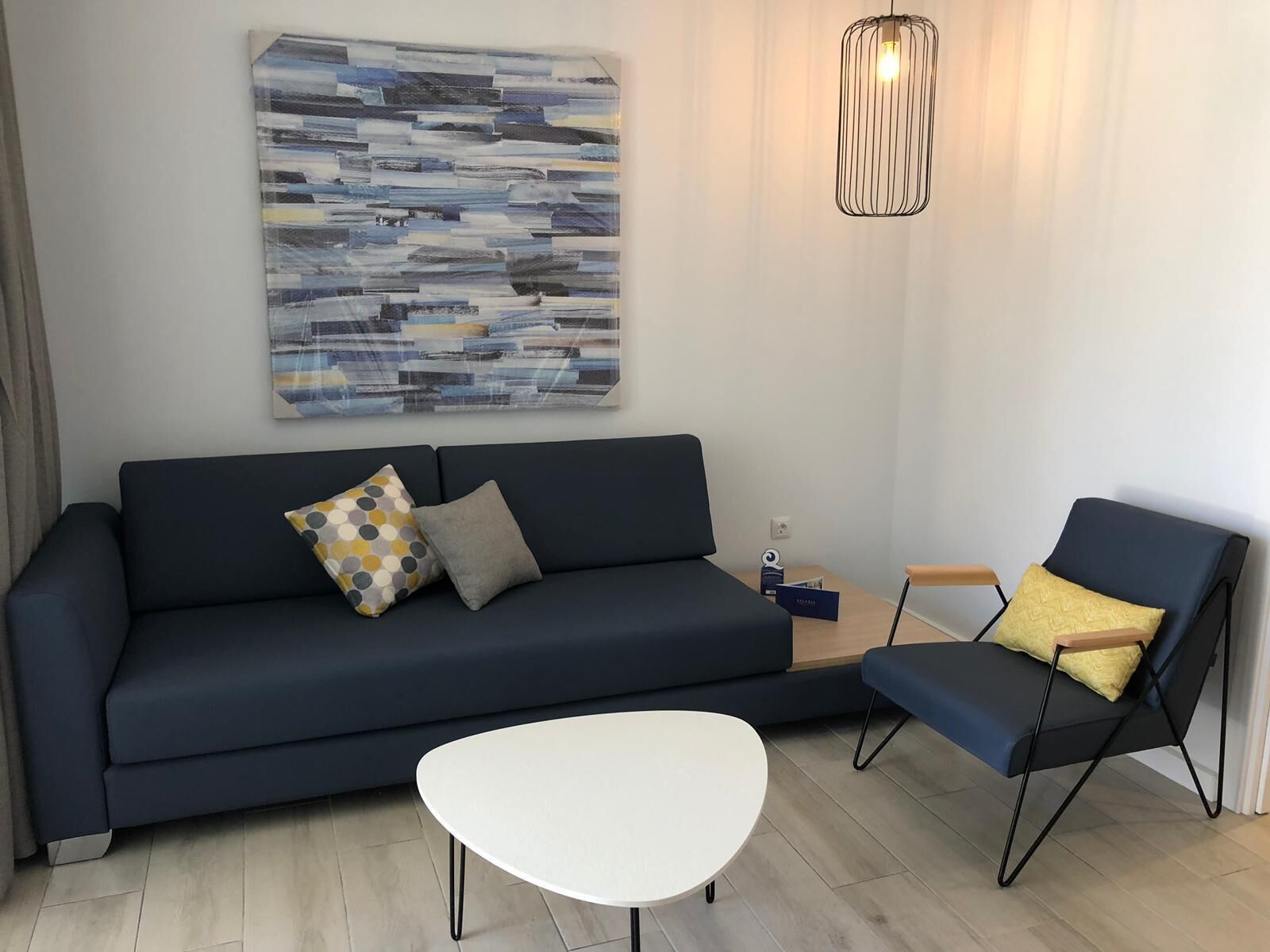 A modern living room with a dark blue sofa, a white coffee table, and a black armchair with a yellow cushion. A contemporary abstract painting hangs on the wall, and a pendant light is visible.