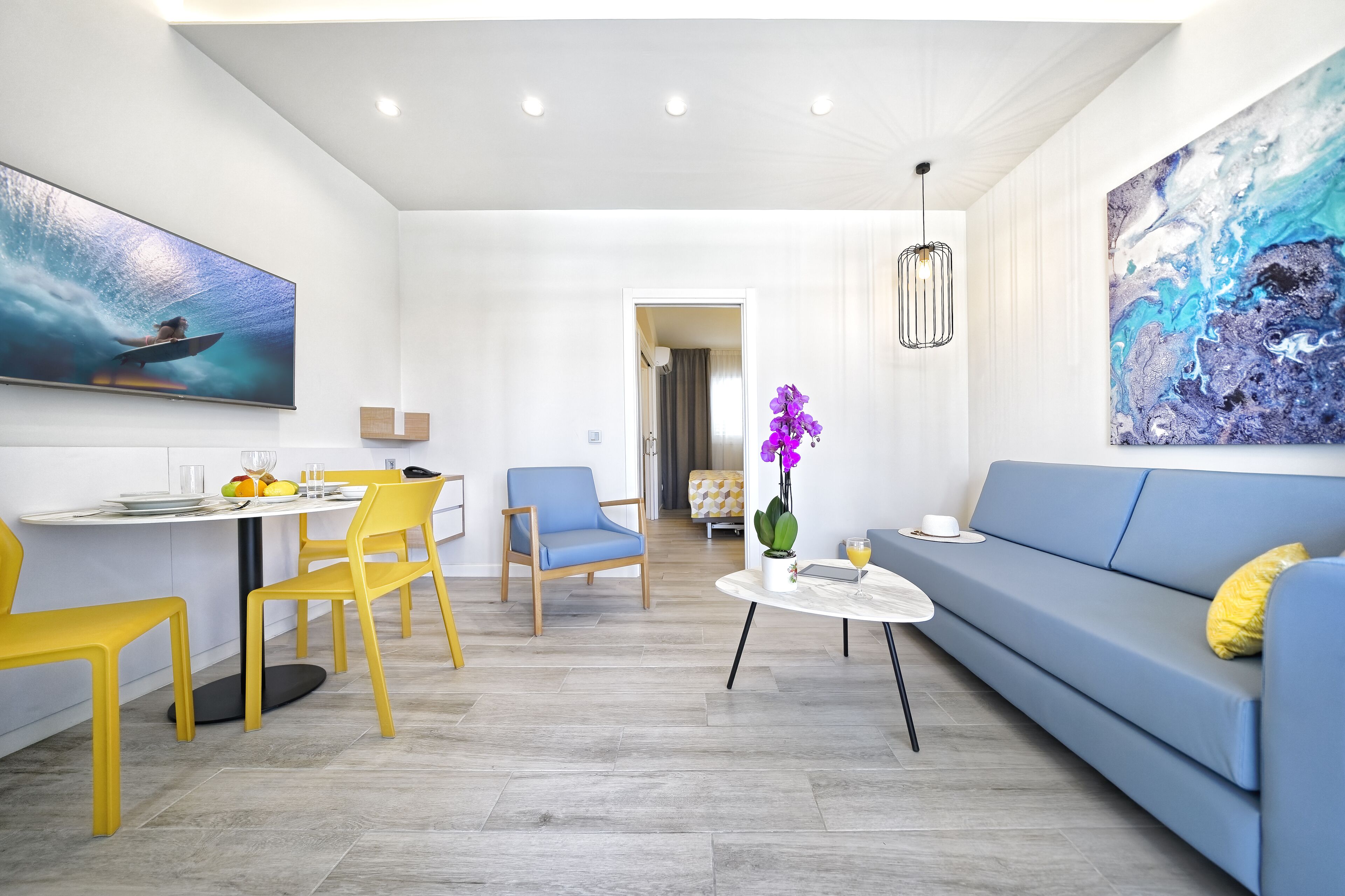 A modern living room with a blue sofa, yellow chairs, a dining table with chairs, a small coffee table, and artwork on the walls.