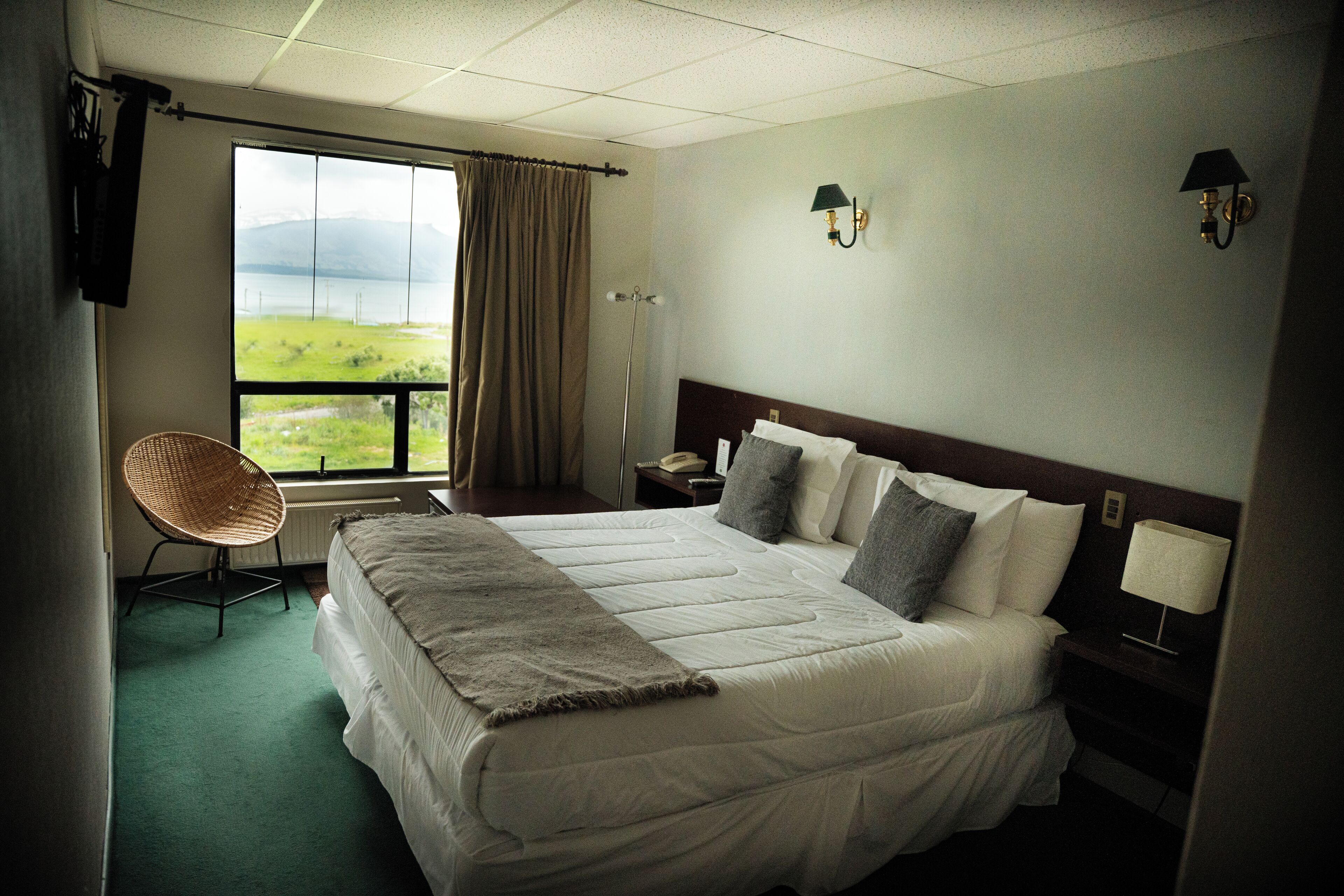 Quarto de hotel com uma cama grande, uma cadeira, janela com vista para o exterior e duas luminárias fixadas na parede.
