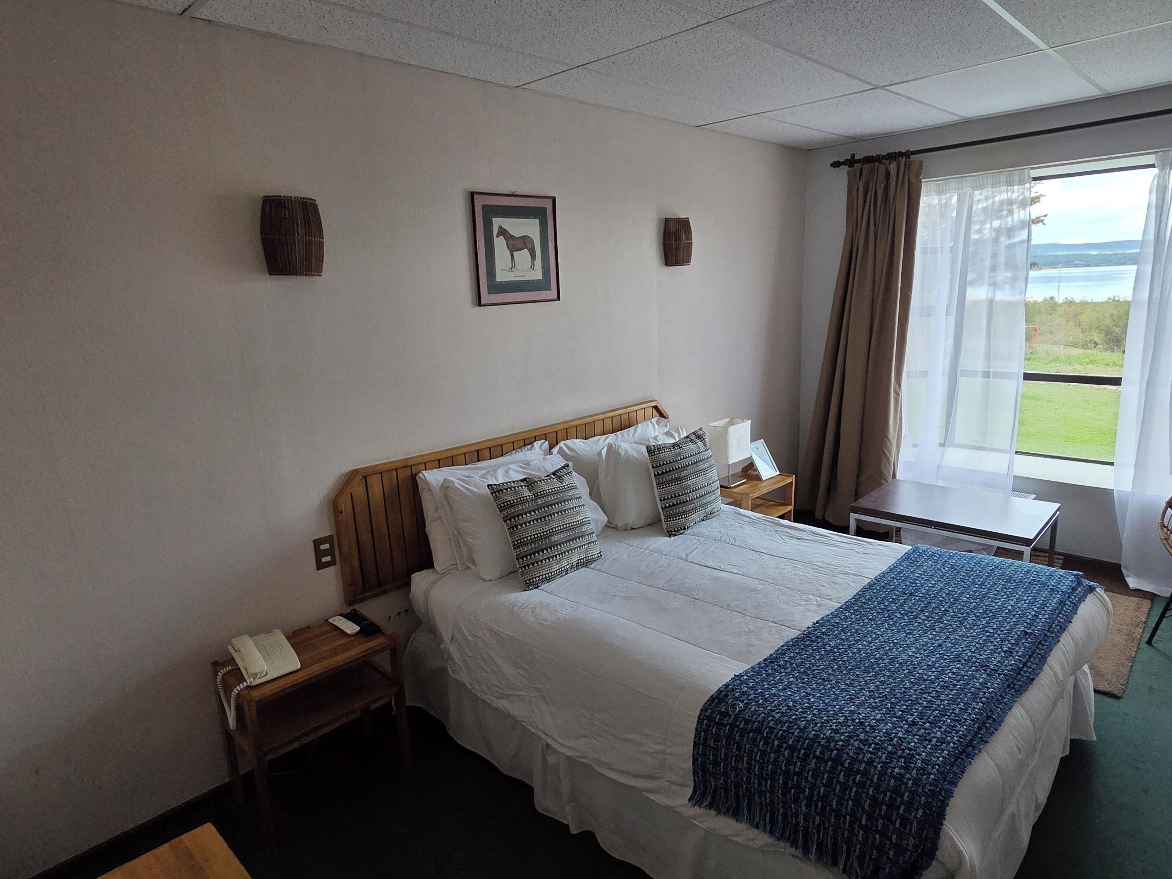 Habitación de hotel con una cama grande, una mesita de noche con lámpara, una pequeña mesa con silla y una ventana con cortinas.