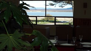 A dining area with a table set for two, a large window with a view of a lake and mountains, and a potted plant in the foreground. - Hotel HD Natales (Natales)