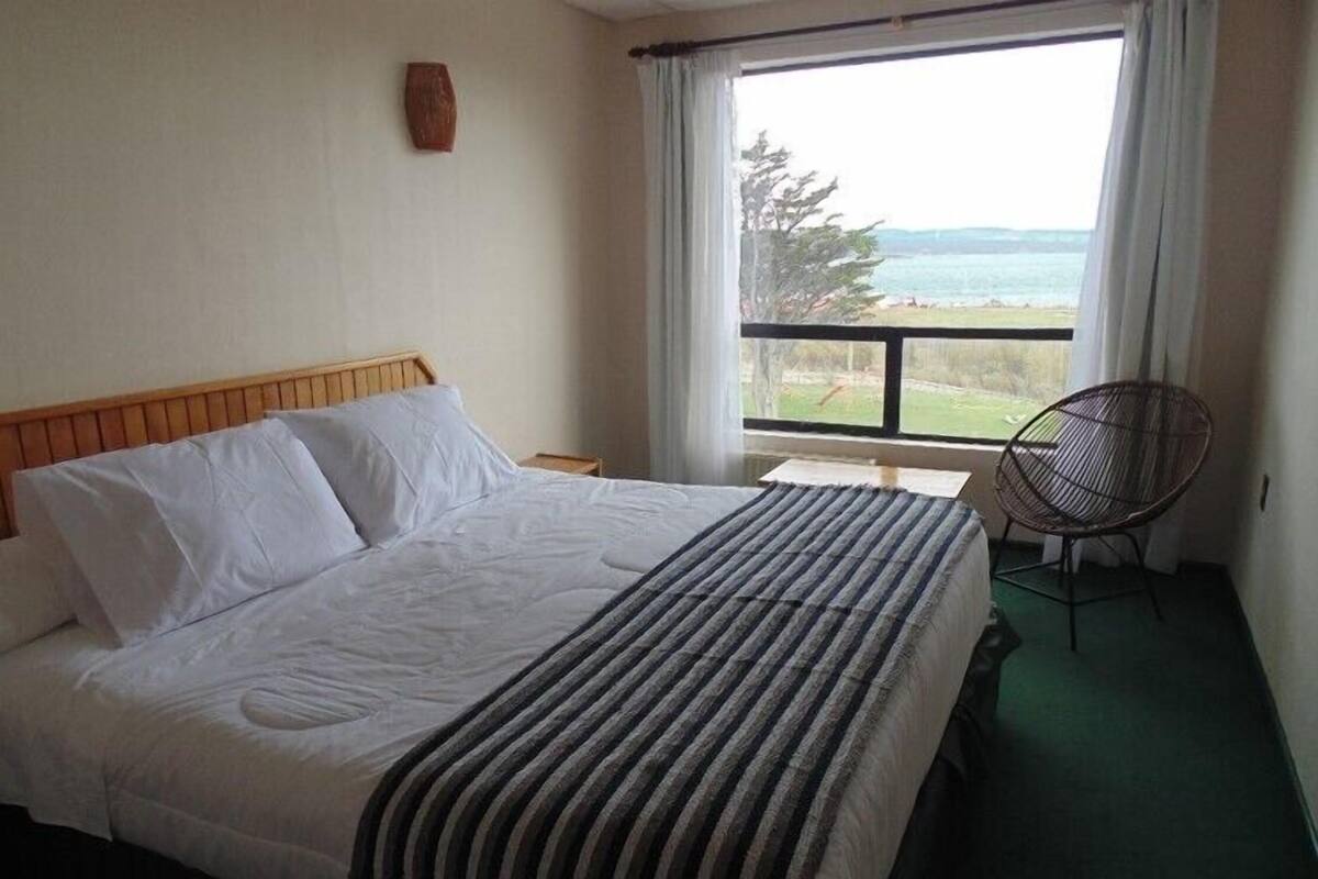 A hotel room with a bed, a window with a view of the sea, a chair, and a wooden headboard.