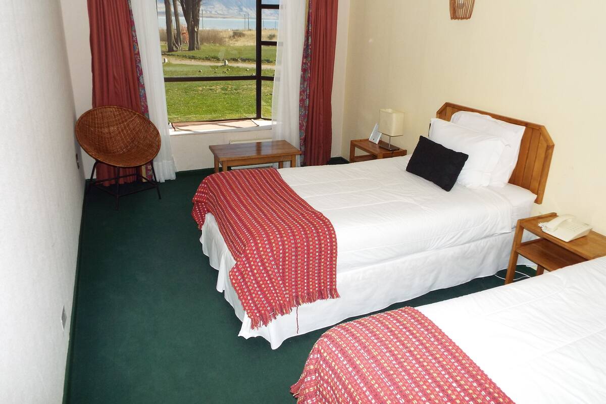 A hotel room with two beds, a chair, a small table, and a view of a lake and mountains.