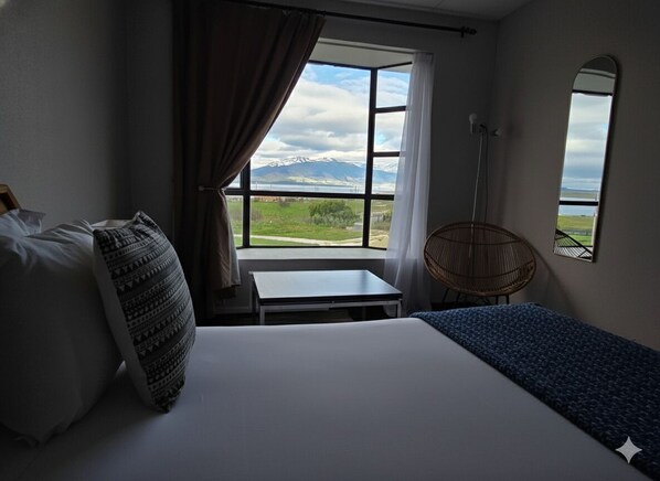 A hotel room with a bed, a small table, a chair, and a window offering a mountain view.