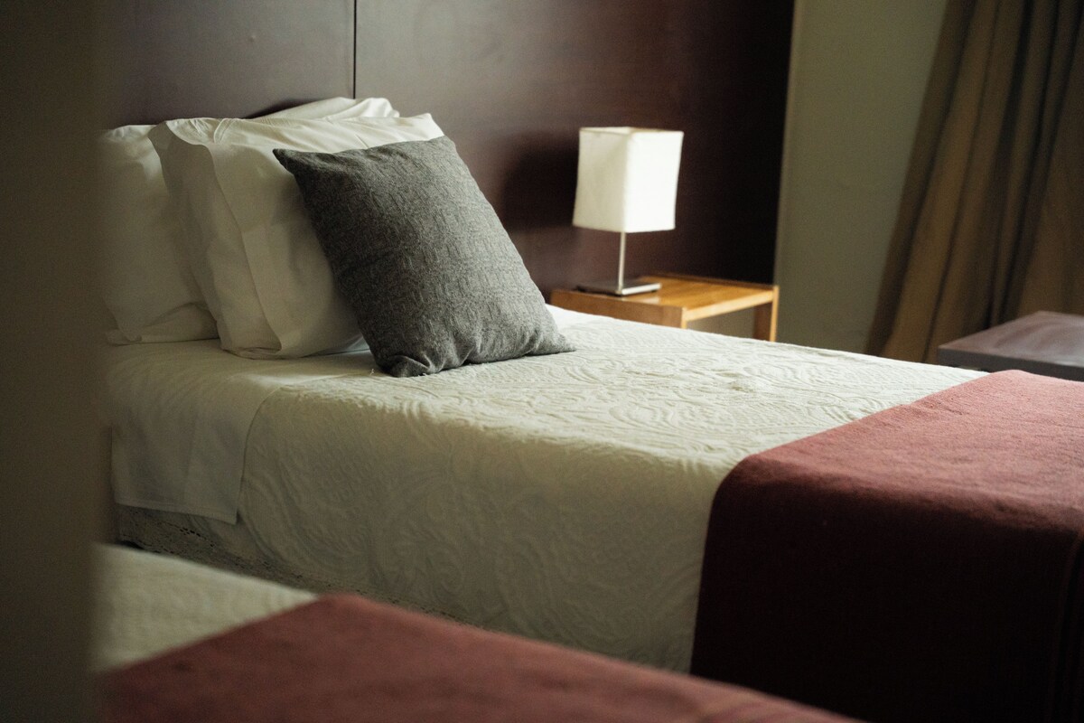 A hotel room with a bed, pillows, a bedside table with a lamp, and a curtain.