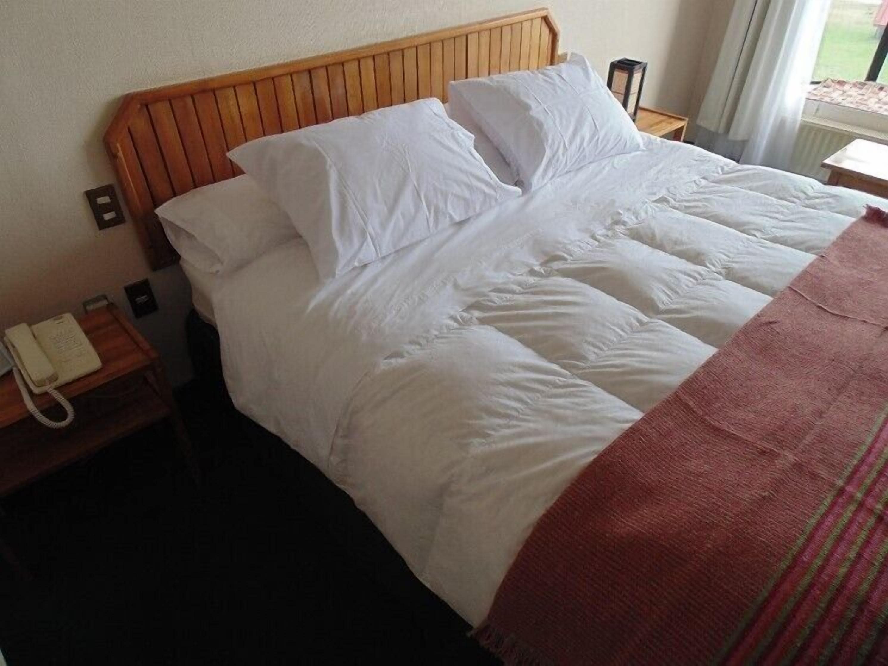 Habitación de hotel con cabecera de madera, ropa de cama blanca, un teléfono en una mesita de noche y una ventana con cortinas.