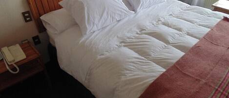 Habitación de hotel con cabecera de madera, ropa de cama blanca, un teléfono en una mesita de noche y una ventana con cortinas.