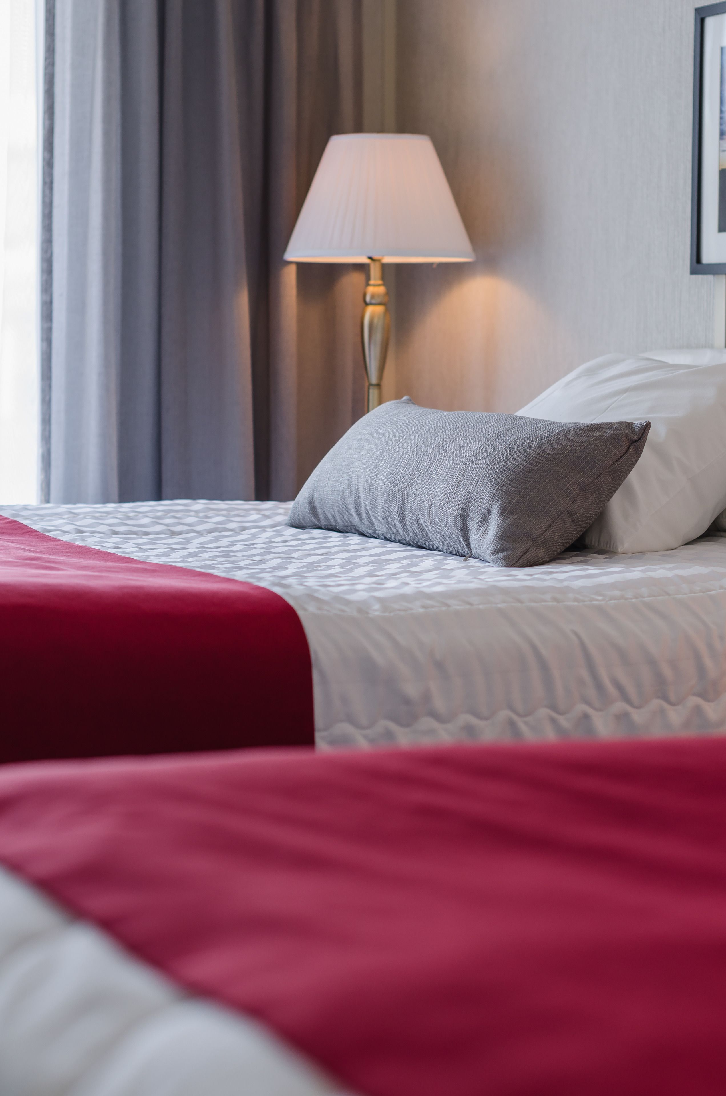 Una cama bien hecha con una colcha roja y un almohadón gris, una lámpara blanca y una cortina gris en una habitación de hotel.