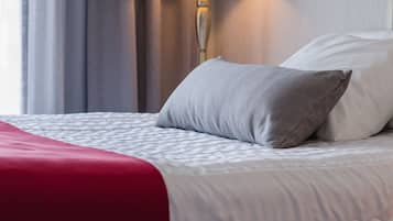 Una cama bien hecha con una colcha roja y un almohadón gris, una lámpara blanca y una cortina gris en una habitación de hotel.