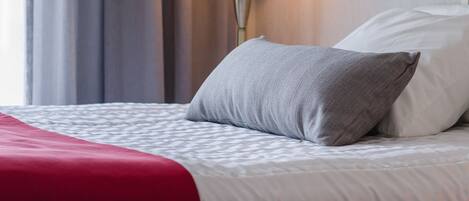 Una cama bien hecha con una colcha roja y un almohadón gris, una lámpara blanca y una cortina gris en una habitación de hotel.