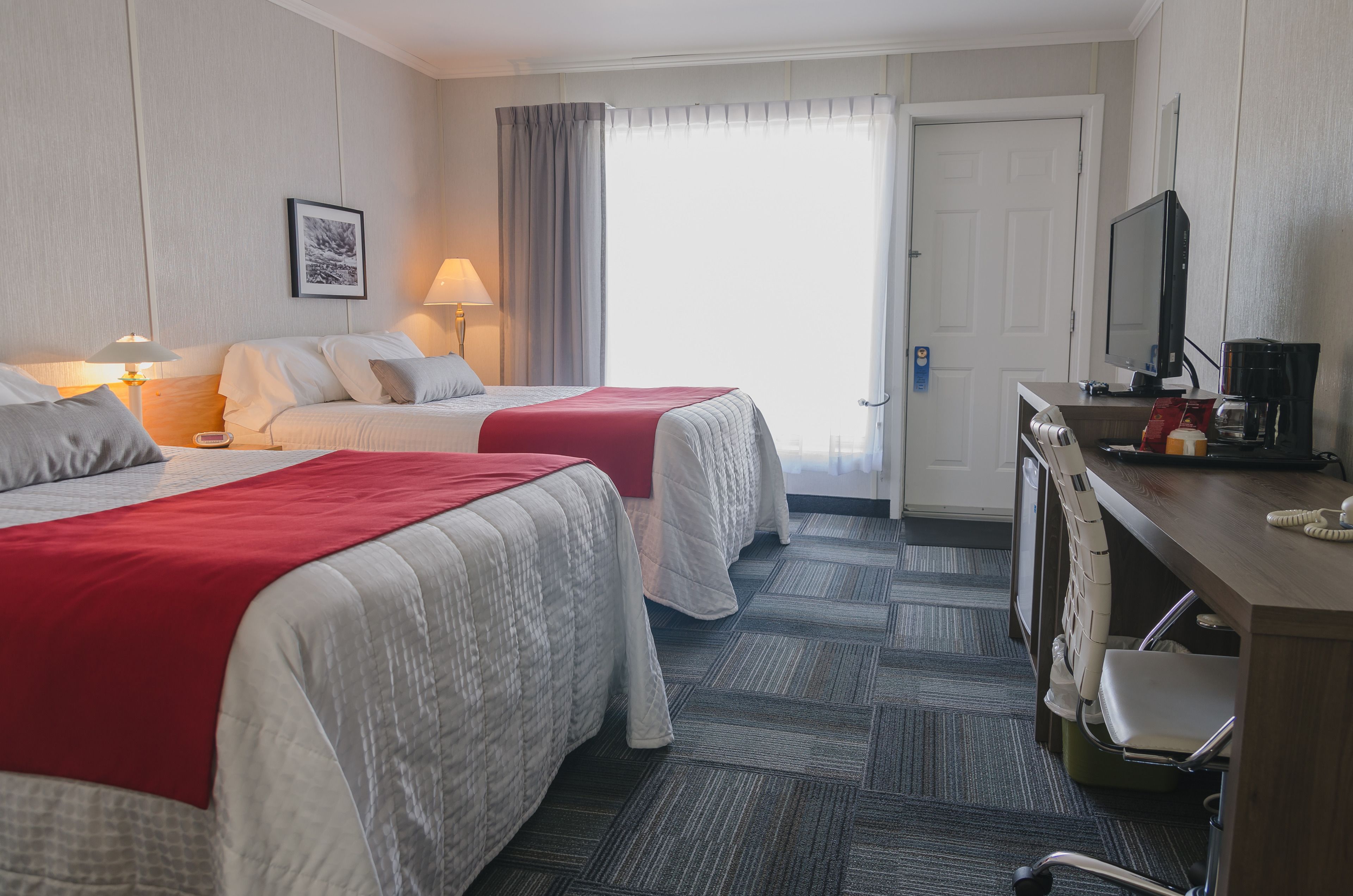 A hotel room with two beds, a desk with a computer and coffee maker, a TV, and a door with a view to the outside.