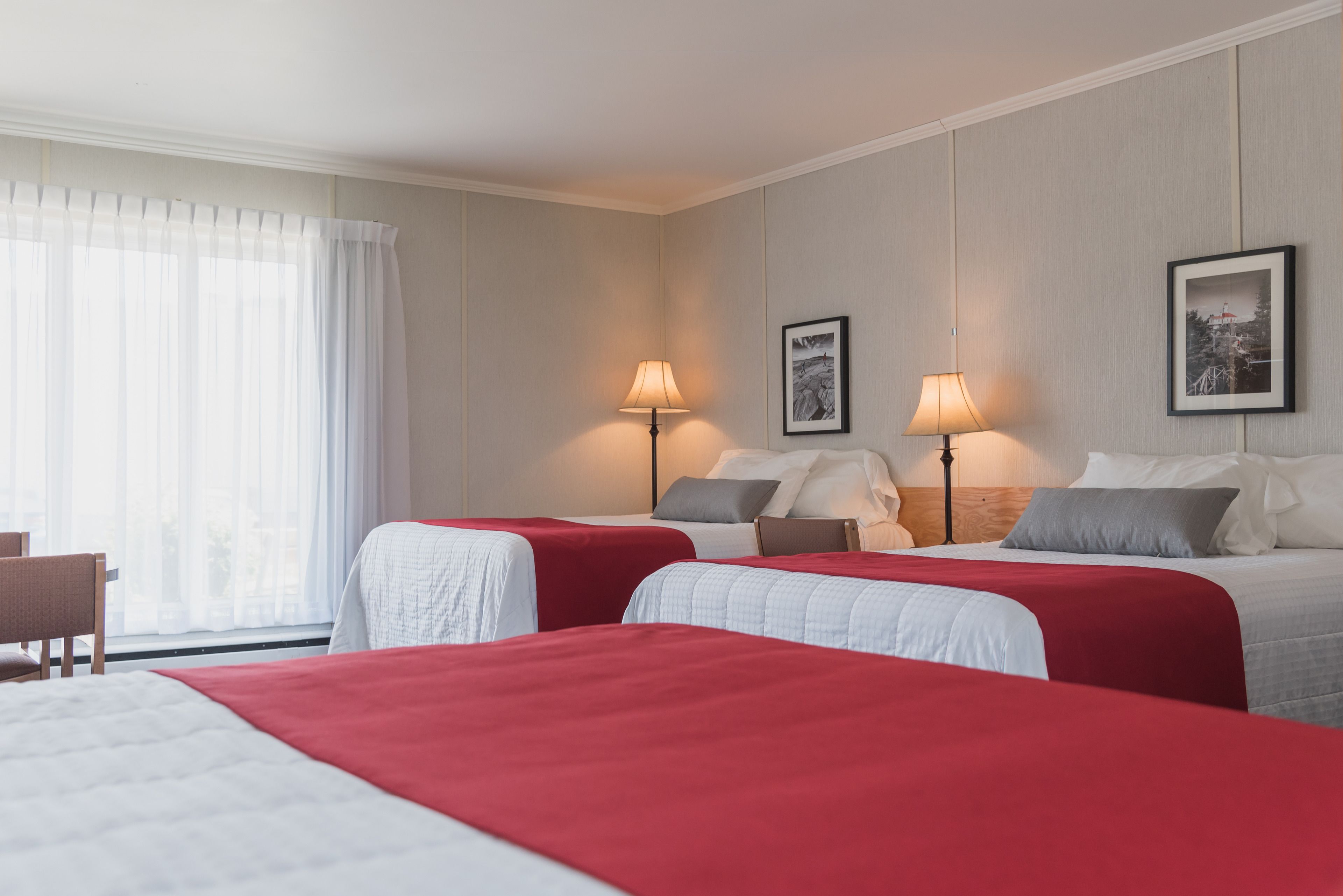 A hotel room with two beds, each with a red bedspread, a wooden headboard, and a nightstand with a lamp. There is a chair and a window with sheer curtains.