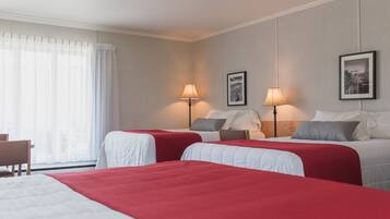 A hotel room with two beds, each with a red bedspread, a wooden headboard, and a nightstand with a lamp. There is a chair and a window with sheer curtains.