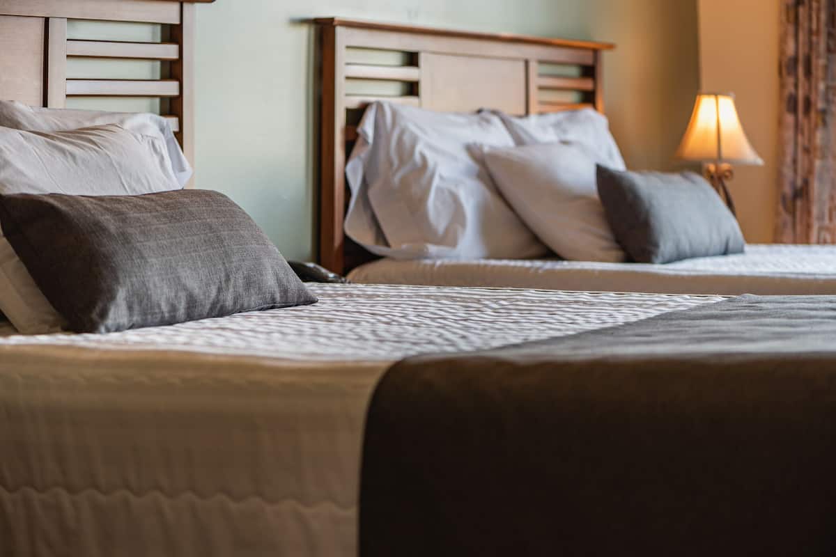 A hotel room with two beds, each with a wooden headboard, white bedding, and a lamp on the nightstand.