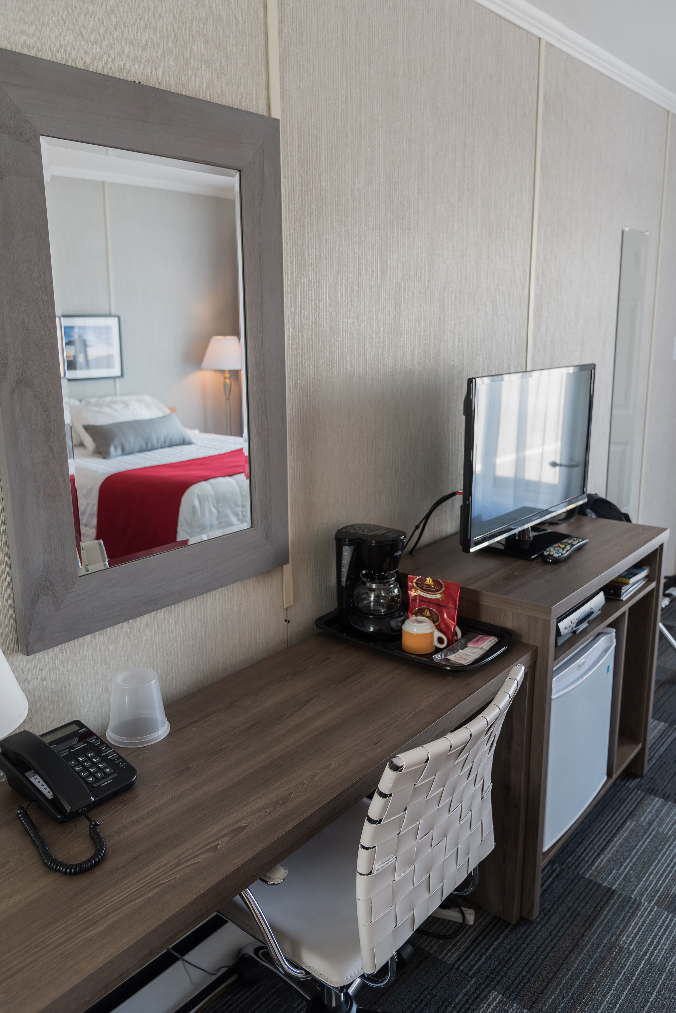 Habitación de hotel con un escritorio de madera, un espejo, televisor, cafetera, teléfono y refrigerador pequeño.