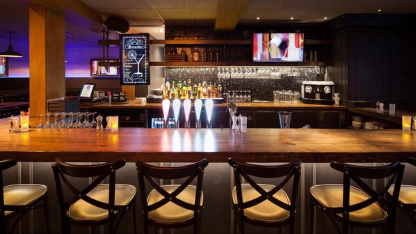 Una zona de bar con una barra alargada, varios taburetes, una pantalla de televisión y una variedad de botellas y vasos en los estantes.