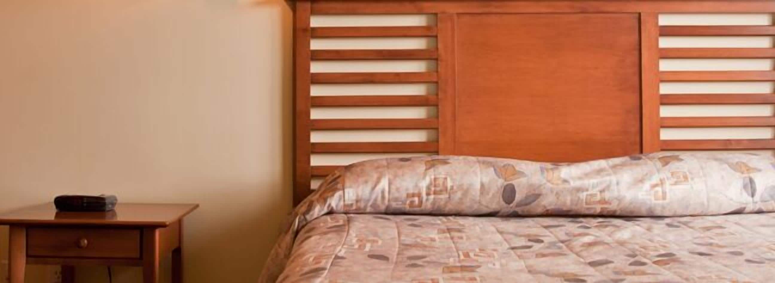 A bedroom with a wooden headboard, a bed with patterned bedding, and a small wooden nightstand with a remote control.