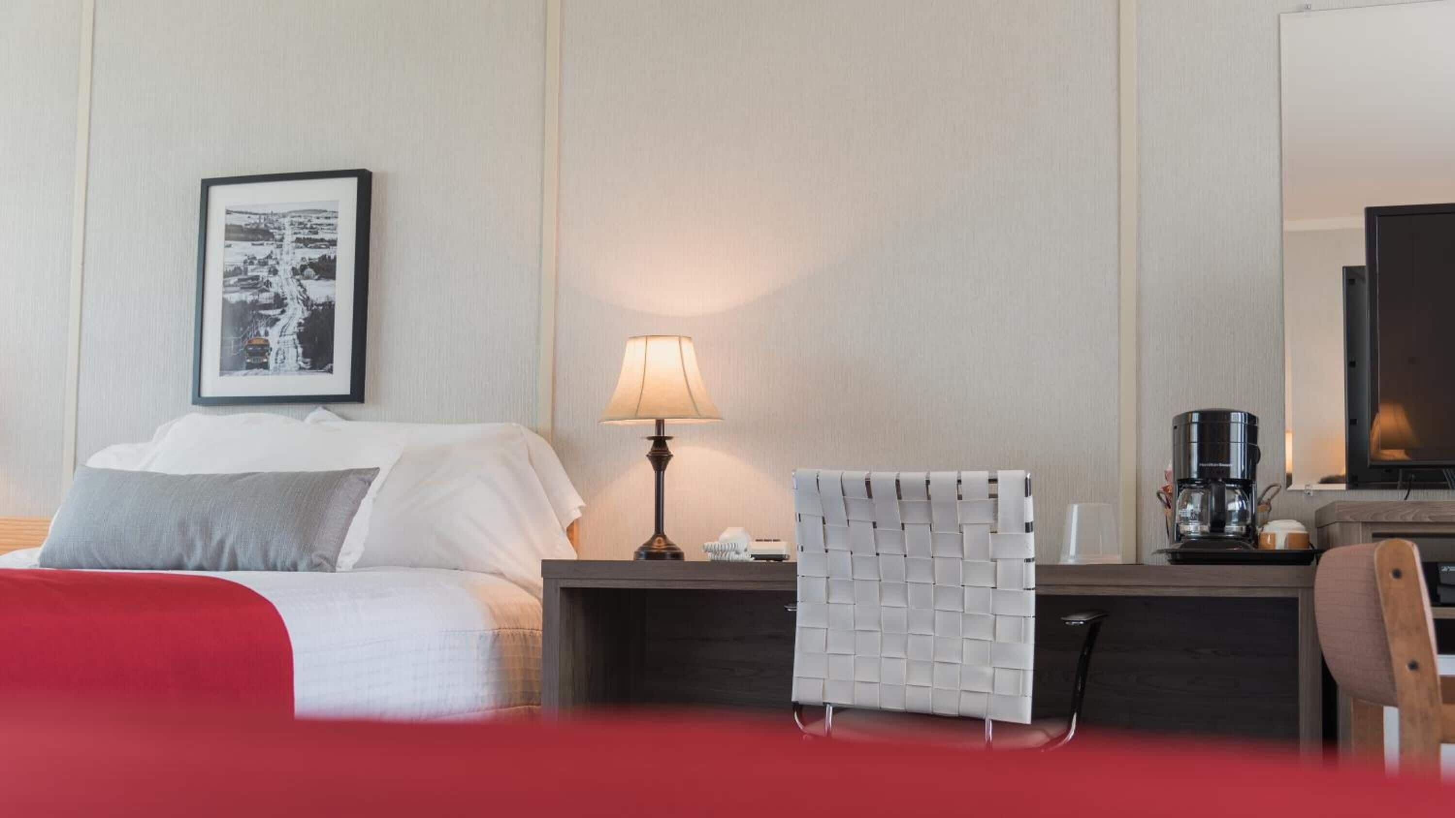 Quarto de hotel com cama, mesa de cabeceira com abajur, uma cadeira, cafeteira e um quadro na parede.