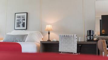 Une chambre d’hôtel avec un lit, une table de chevet avec une lampe, une chaise, une machine à café et un tableau encadré au mur.