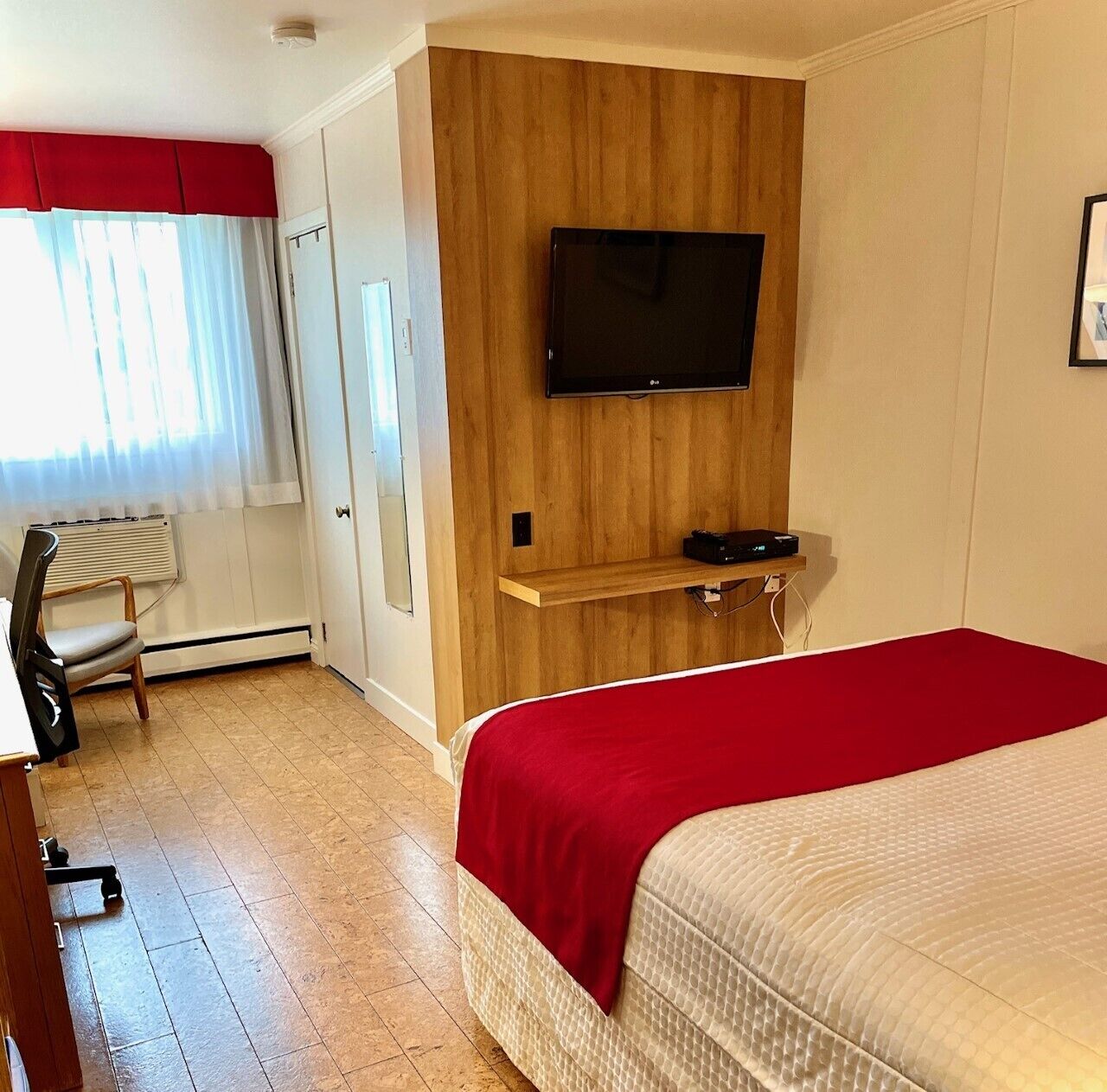 A hotel room with a bed, a TV mounted on a wooden panel wall, a chair, and a radiator.