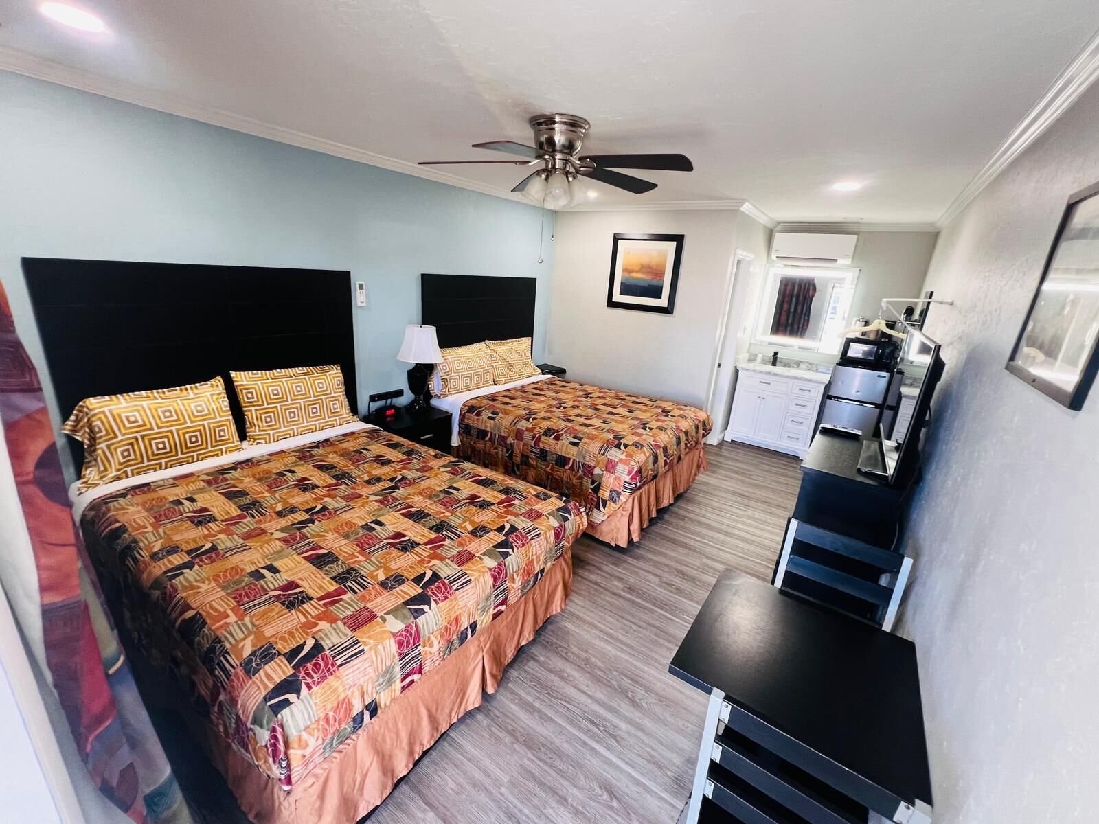 A hotel room with two beds, a ceiling fan, a desk, and a kitchenette.
