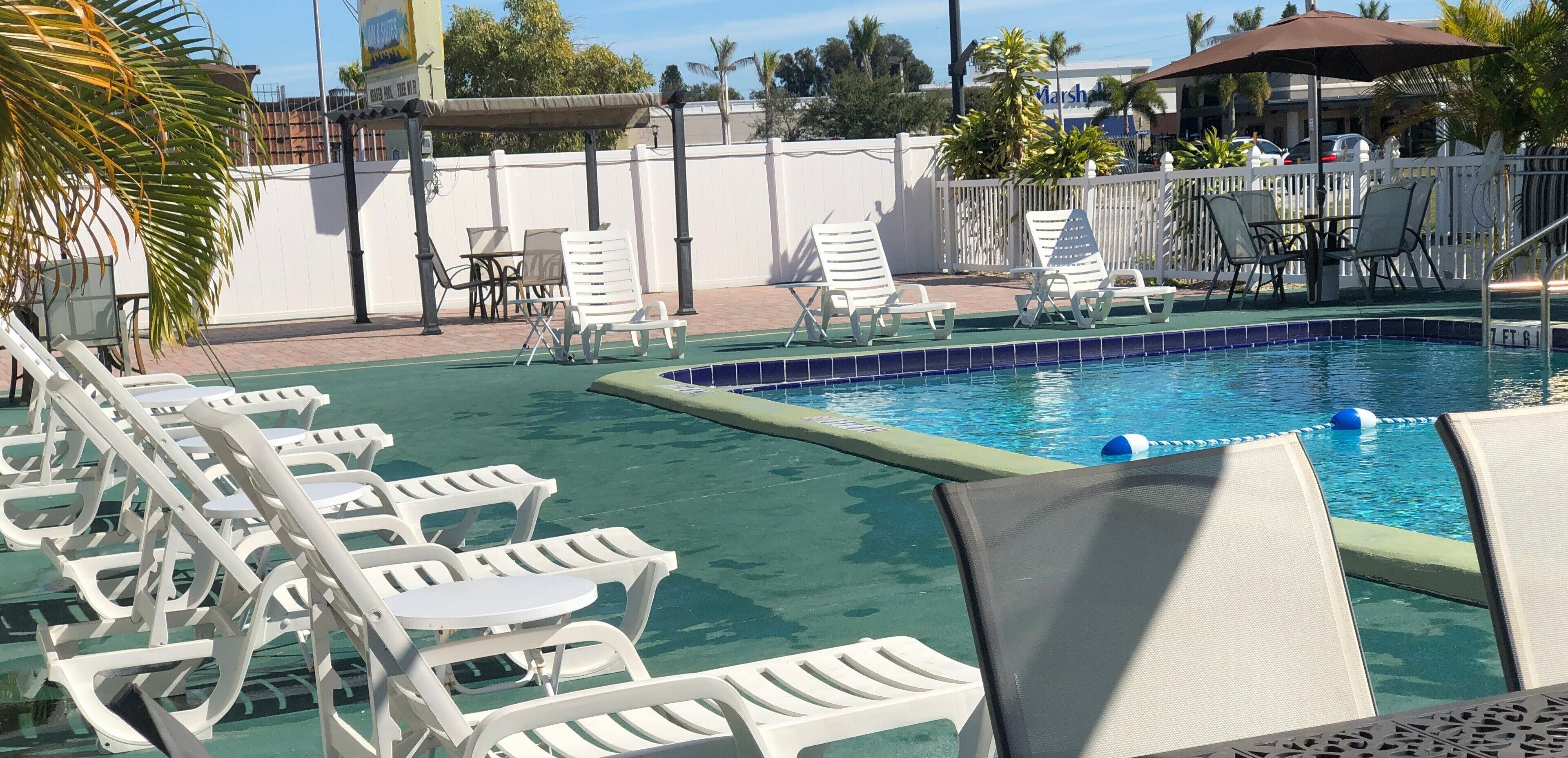 Zona de piscina con sillones blancos y una pequeña mesa, rodeada por una valla blanca y palmeras.