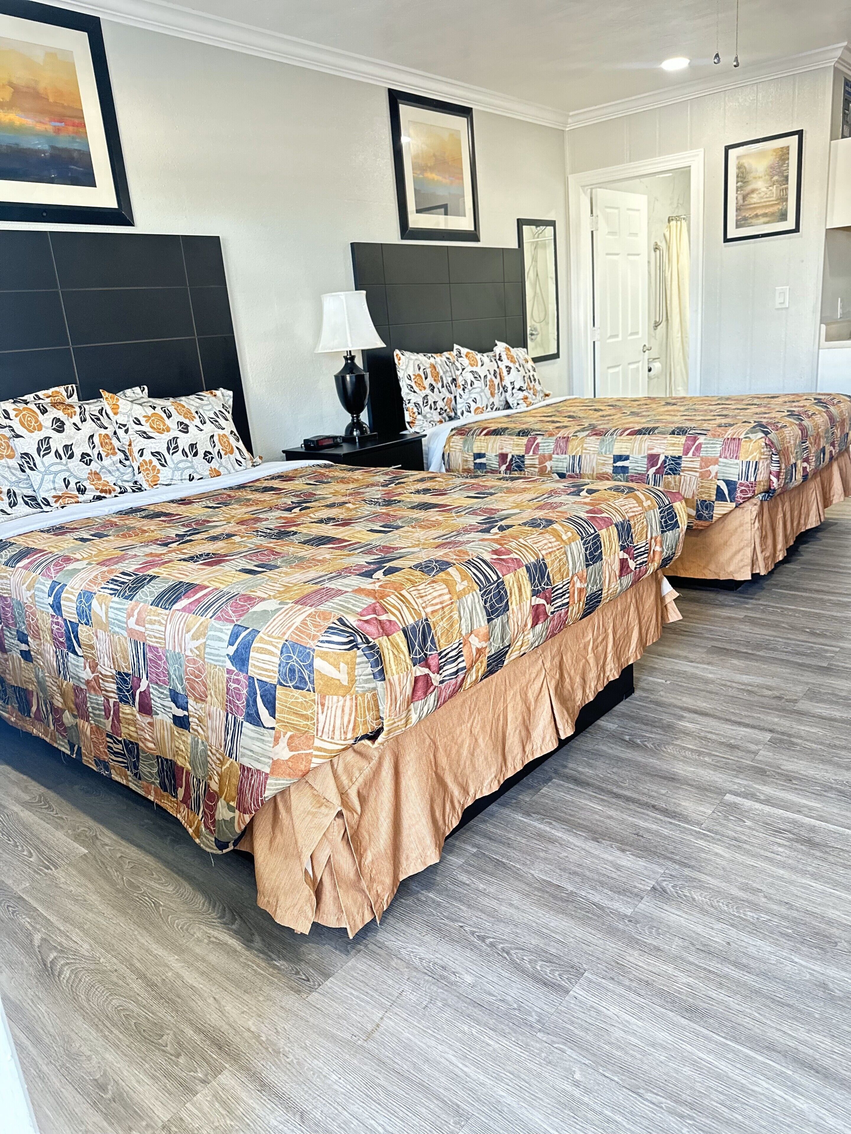 Habitación de hotel con dos camas, cada una con una colcha estampada y almohadas a juego. Hay una mesita de noche con una lámpara, una puerta al baño y cuadros en las paredes.