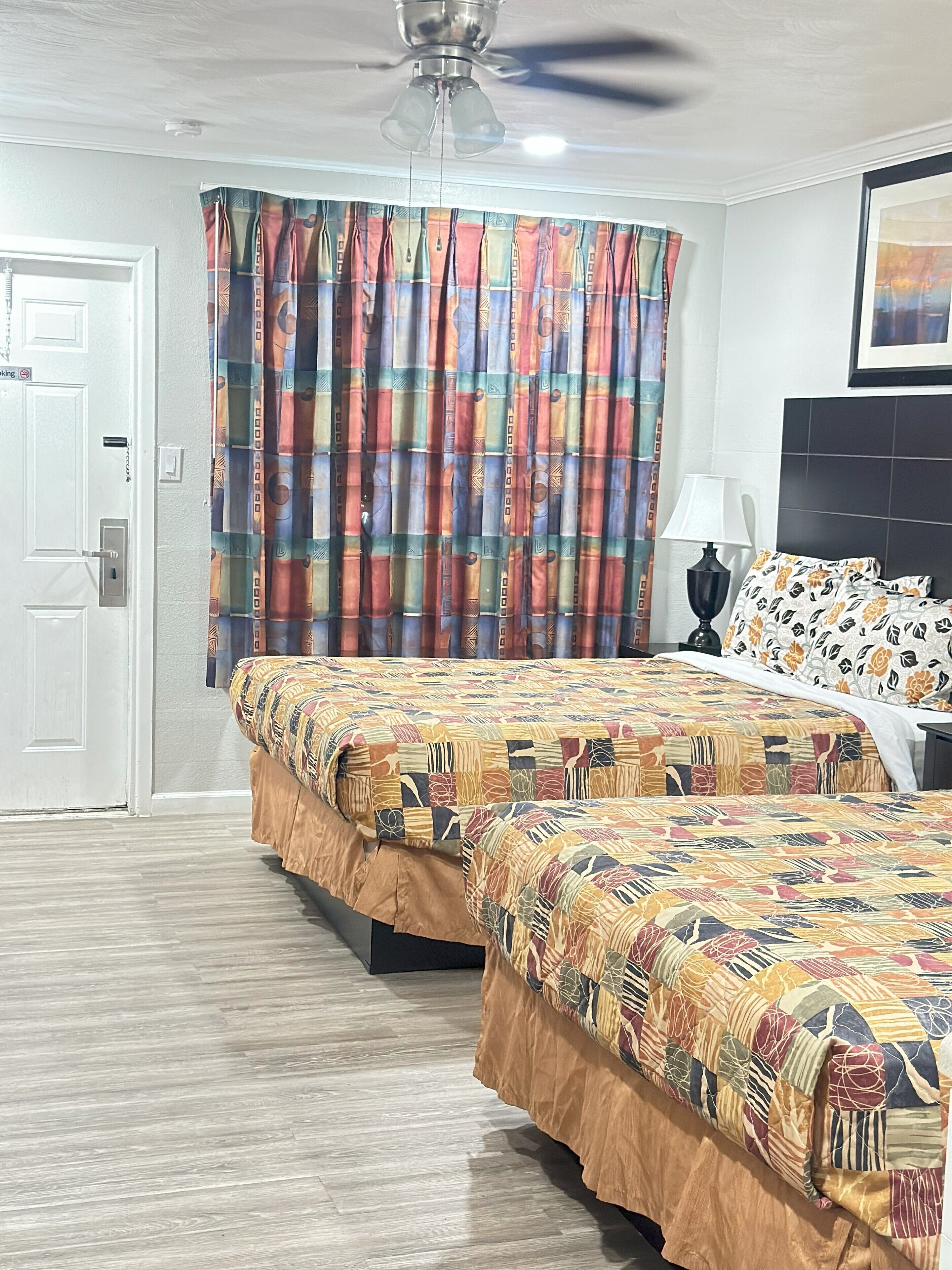 A hotel room with two beds, a ceiling fan, a window with patterned curtains, and a framed picture on the wall.