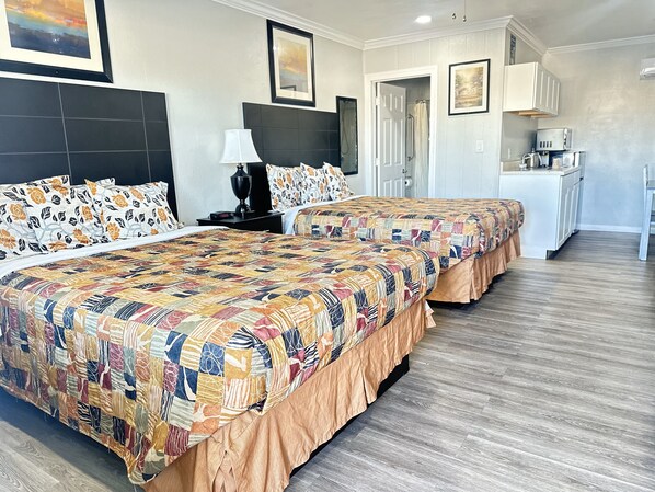 A hotel room with two beds, a nightstand, a lamp, a bathroom door, and a kitchenette.