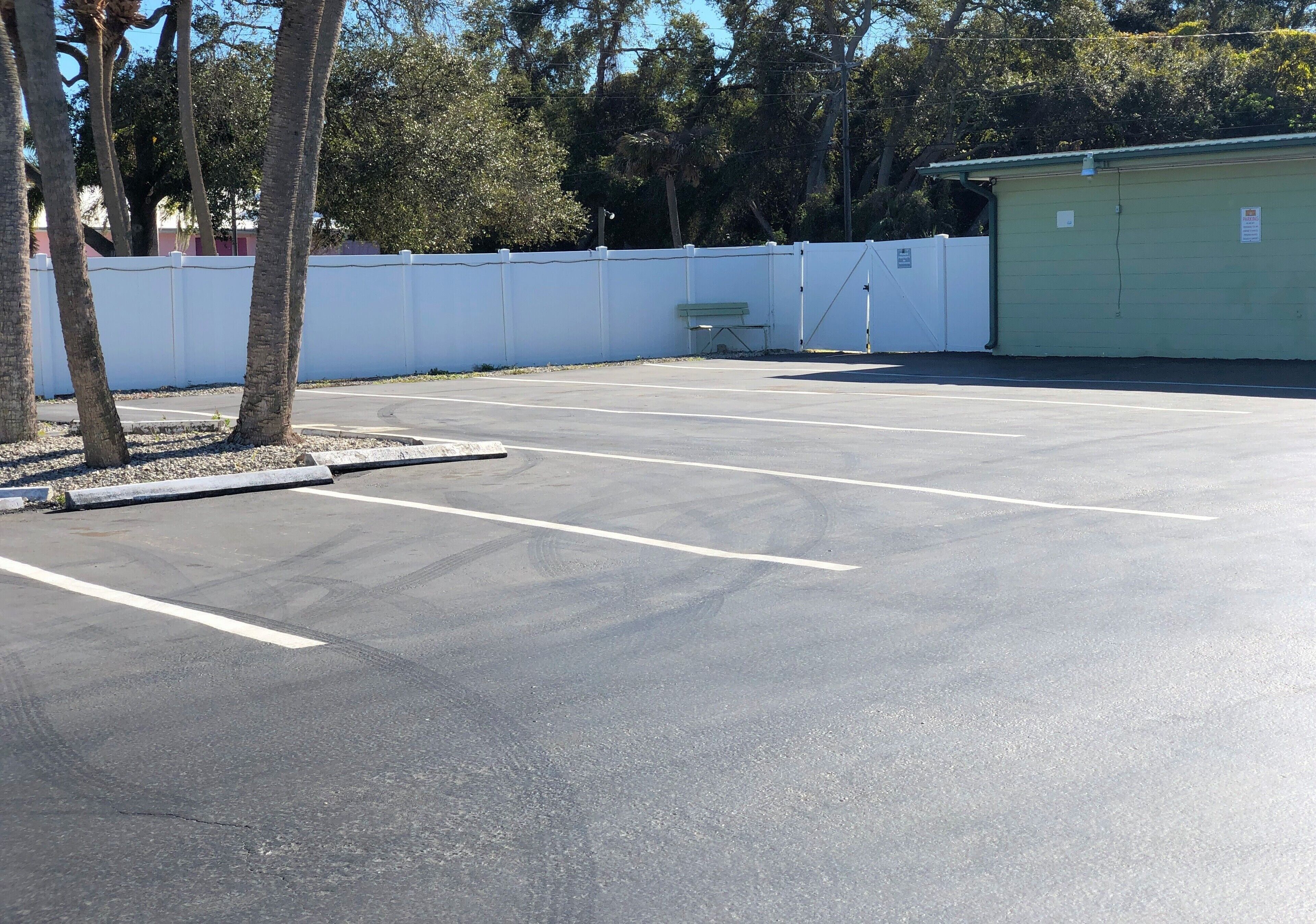 Un estacionamiento con líneas blancas que delimitan los espacios, un edificio verde y una cerca blanca.