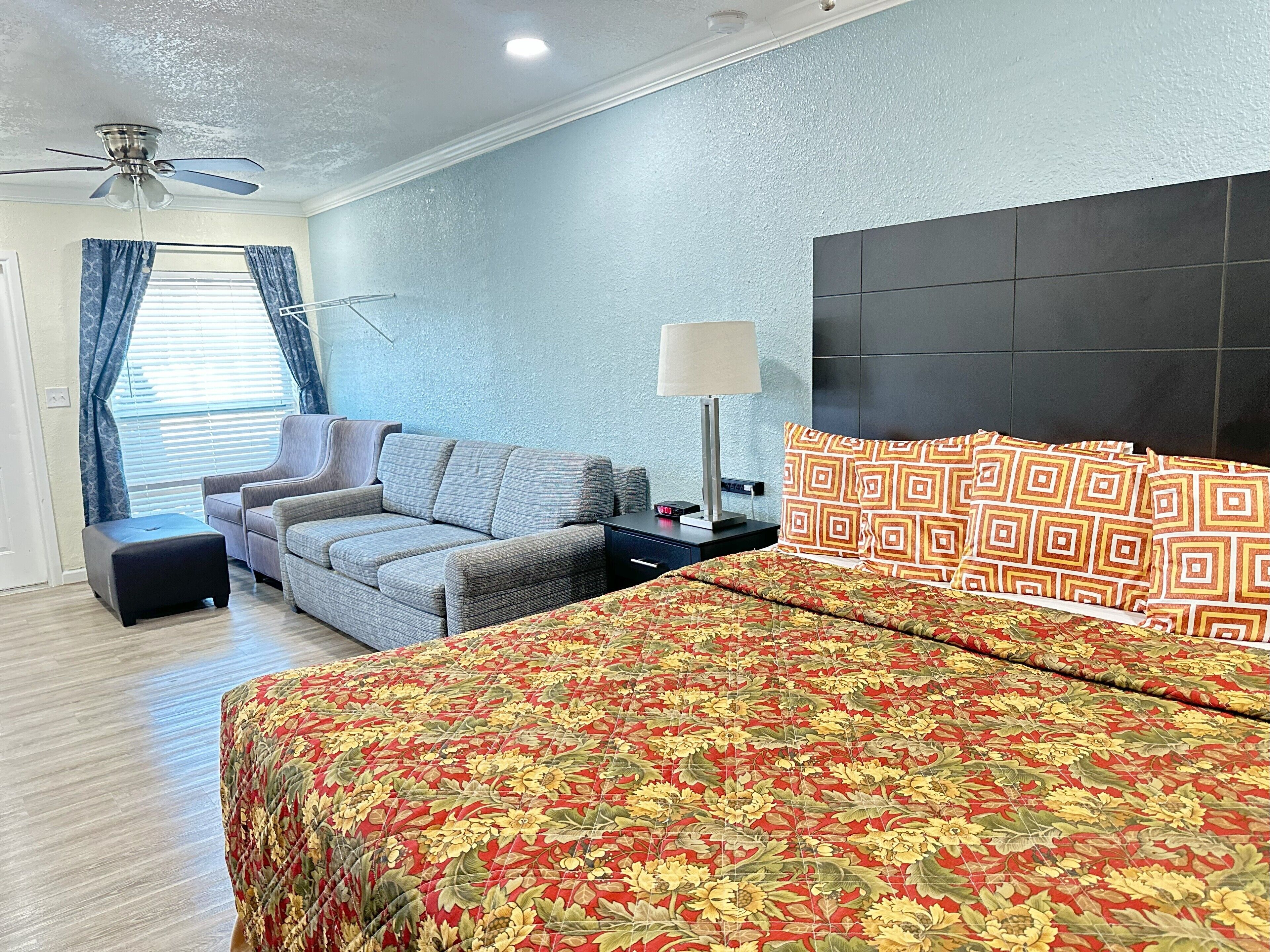 A hotel room with a large bed, a sofa, a small table, a lamp, and a ceiling fan.