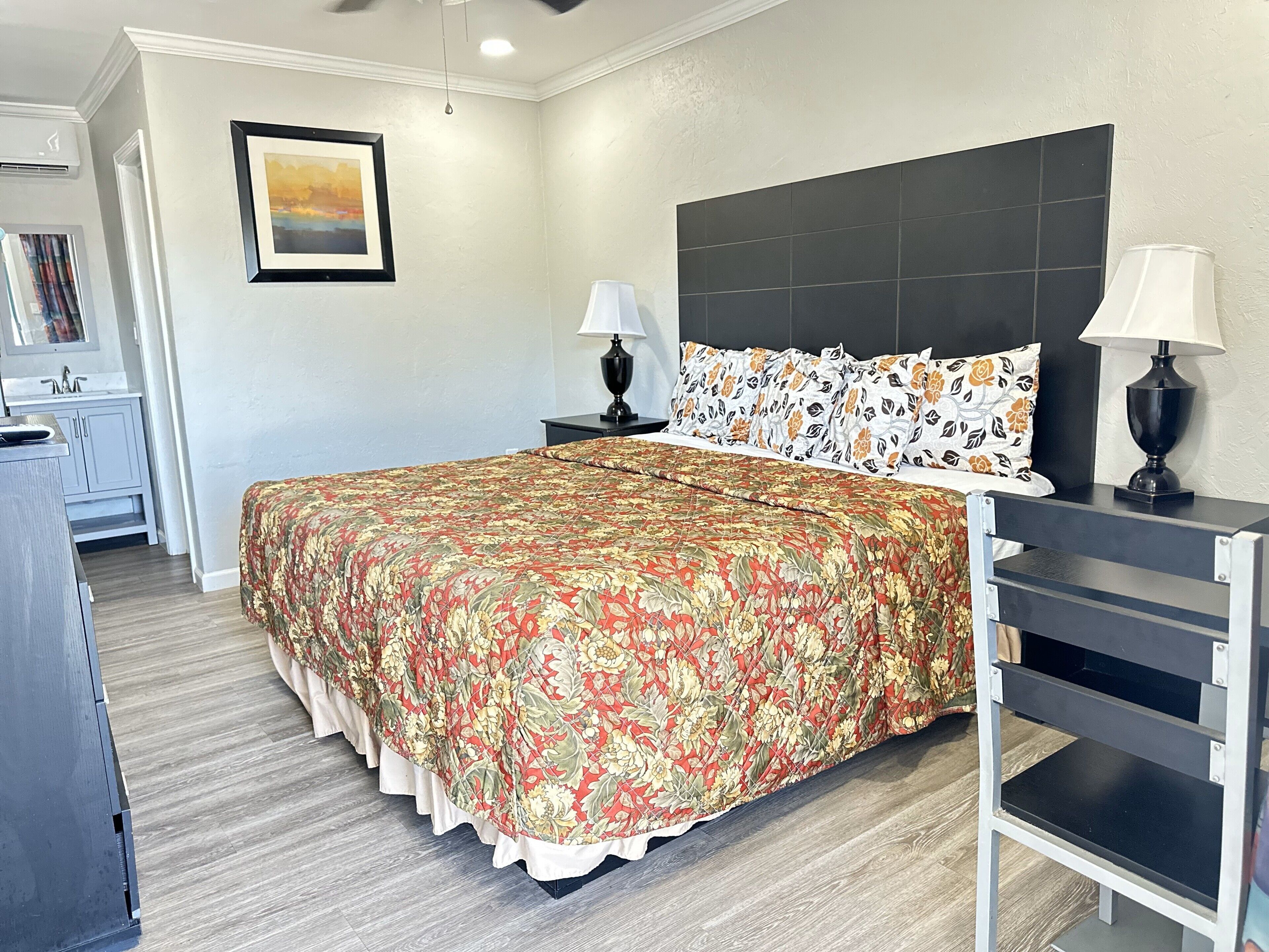 Une chambre à coucher avec un grand lit, deux tables de chevet avec des lampes, une commode, un tableau encadré au mur et un ventilateur de plafond.