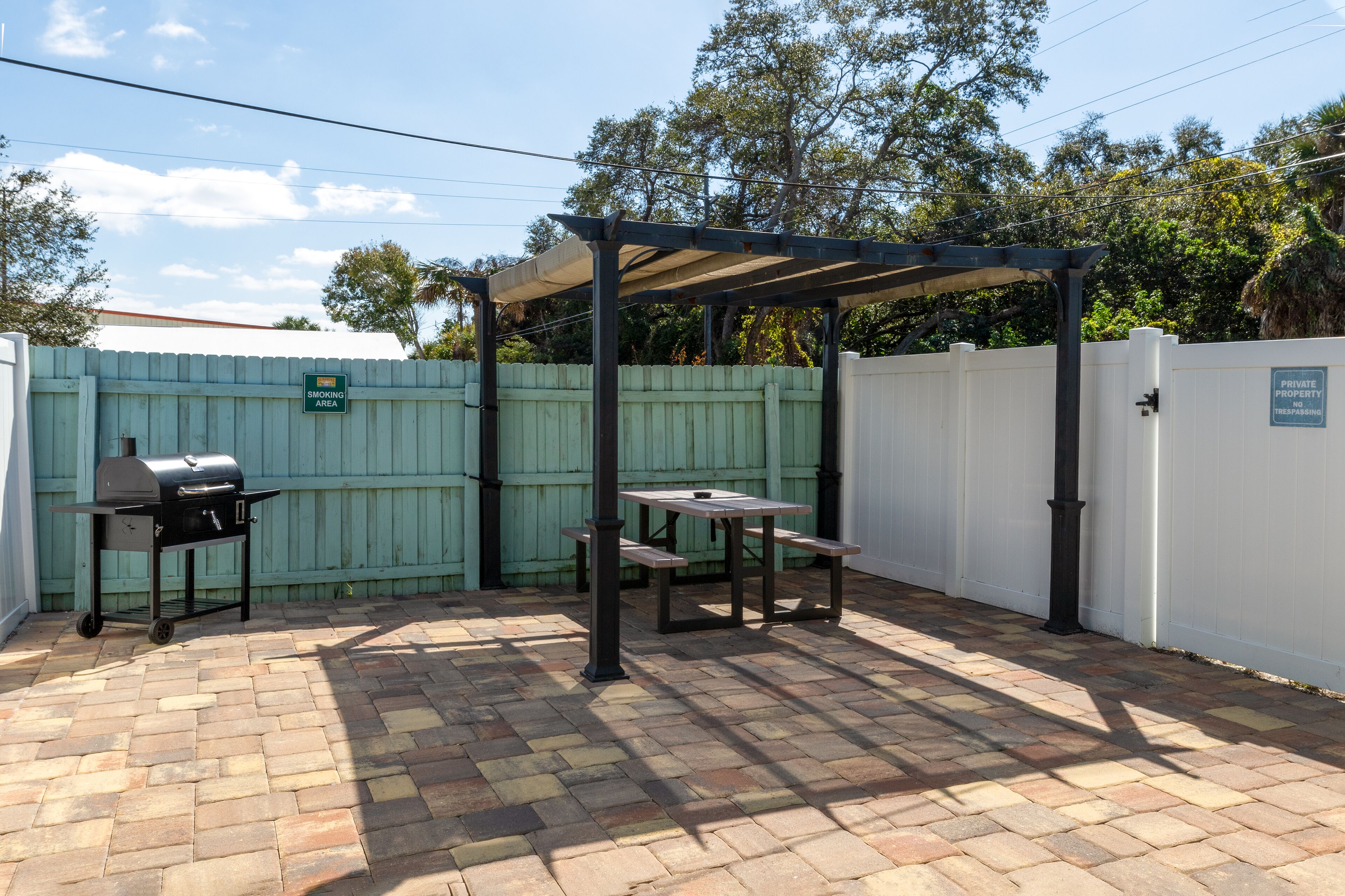Une terrasse équipée d’un barbecue, d’une table de pique-nique et d’un espace salon abrité, le tout délimité par une clôture blanche.