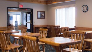 A dining area with wooden tables and chairs, an exit sign, a clock on the wall, and a window with blinds. - White Oak Inn & Suites (Deer River)