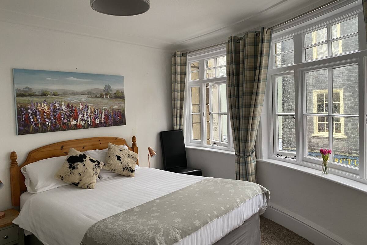 A bedroom with a bed, a painting, a window with curtains, and a desk.