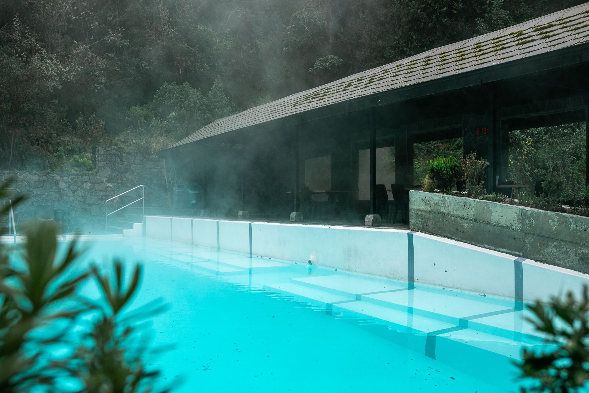 A clear blue pool with a stone wall and a covered area with benches.