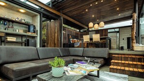 Uma sala de estar moderna com um amplo sofá modular, uma mesa de centro com livros e uma planta, e uma área de cozinha com armários de madeira e uma luminária pendente.