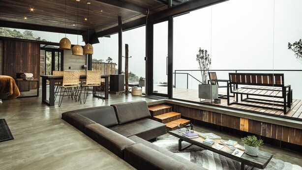 Ampla sala de estar com um sofá grande, mesa de jantar e cadeiras, varanda com deck de madeira e janelas grandes com vista para o exterior.