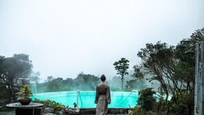 Uma pessoa caminhando em direção a uma piscina sob uma atmosfera nebulosa, cercada por elementos naturais e um caminho de pedra.
