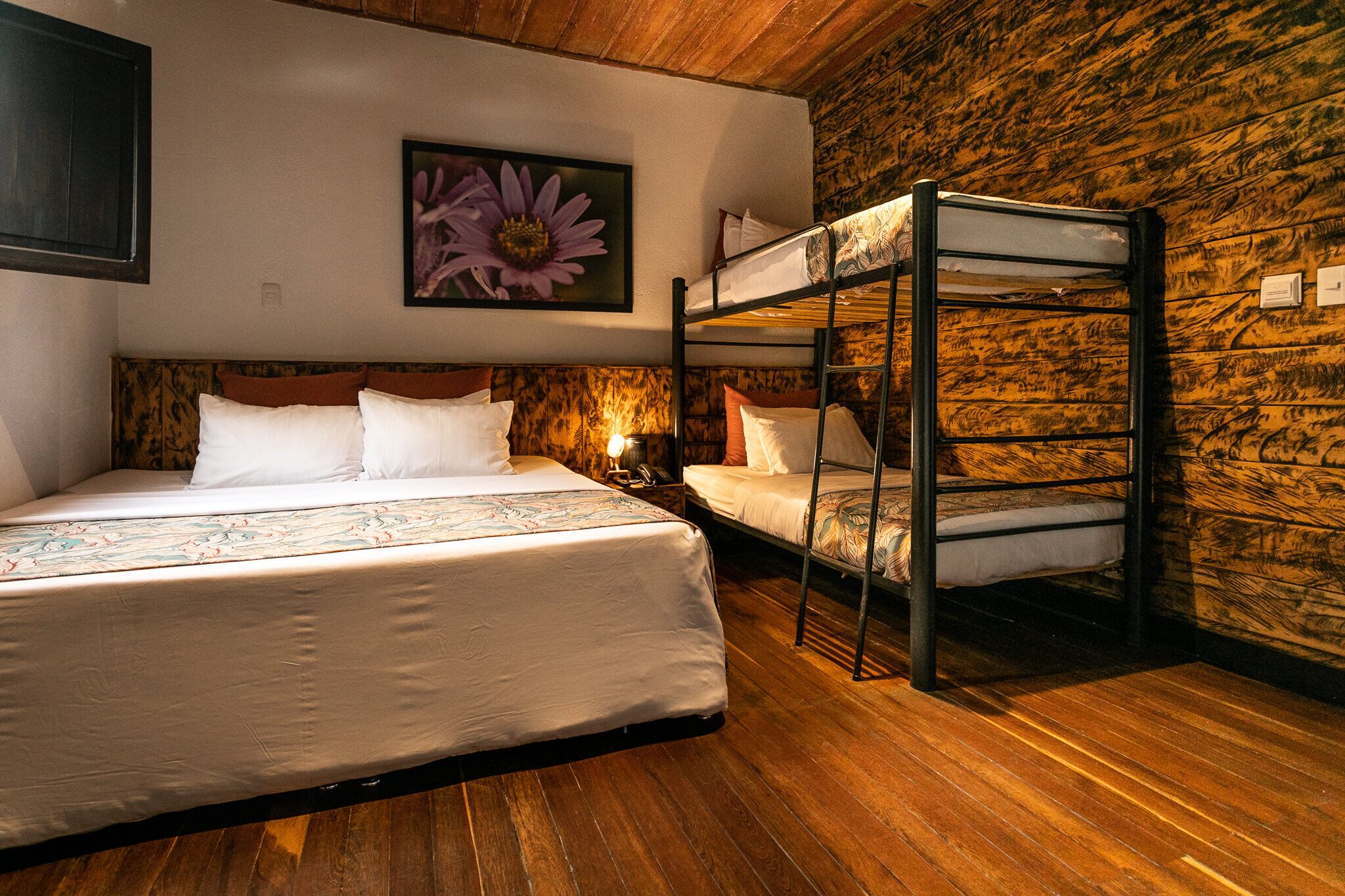 A room with a bunk bed, a large bed with a floral bedspread, a wooden wall, and a framed flower painting.
