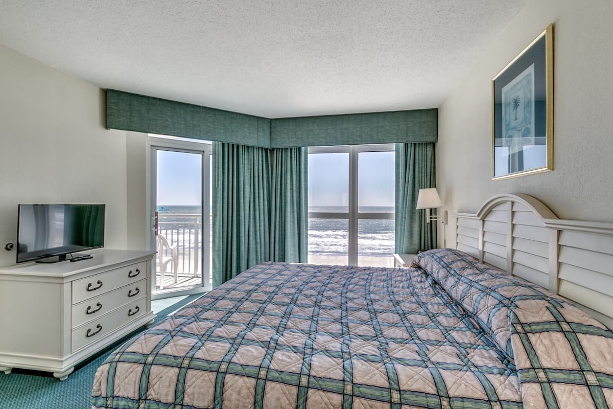A hotel room with a large bed, a TV on a dresser, and a balcony with a view of the ocean.