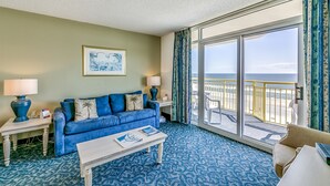 A room with a blue sofa, two lamps, a coffee table, and a large window with a view of the beach. - Watch Resort by Condo-World (North Myrtle Beach)