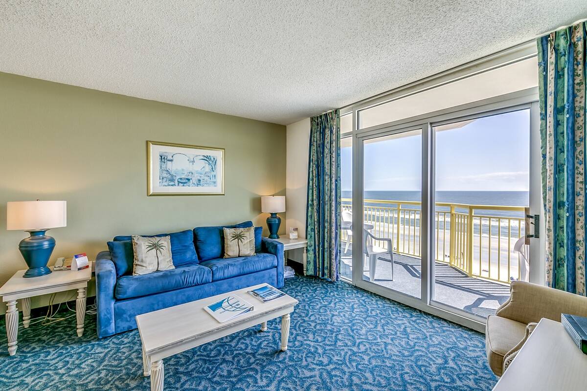 A room with a blue sofa, two lamps, a coffee table, and a large window with a view of the beach.