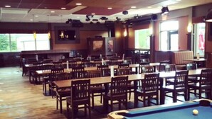 Une table de billard avec une queue et une boule, entourée de plusieurs tables et chaises en bois, dans une pièce dotée de fenêtres et d’un éclairage au plafond.