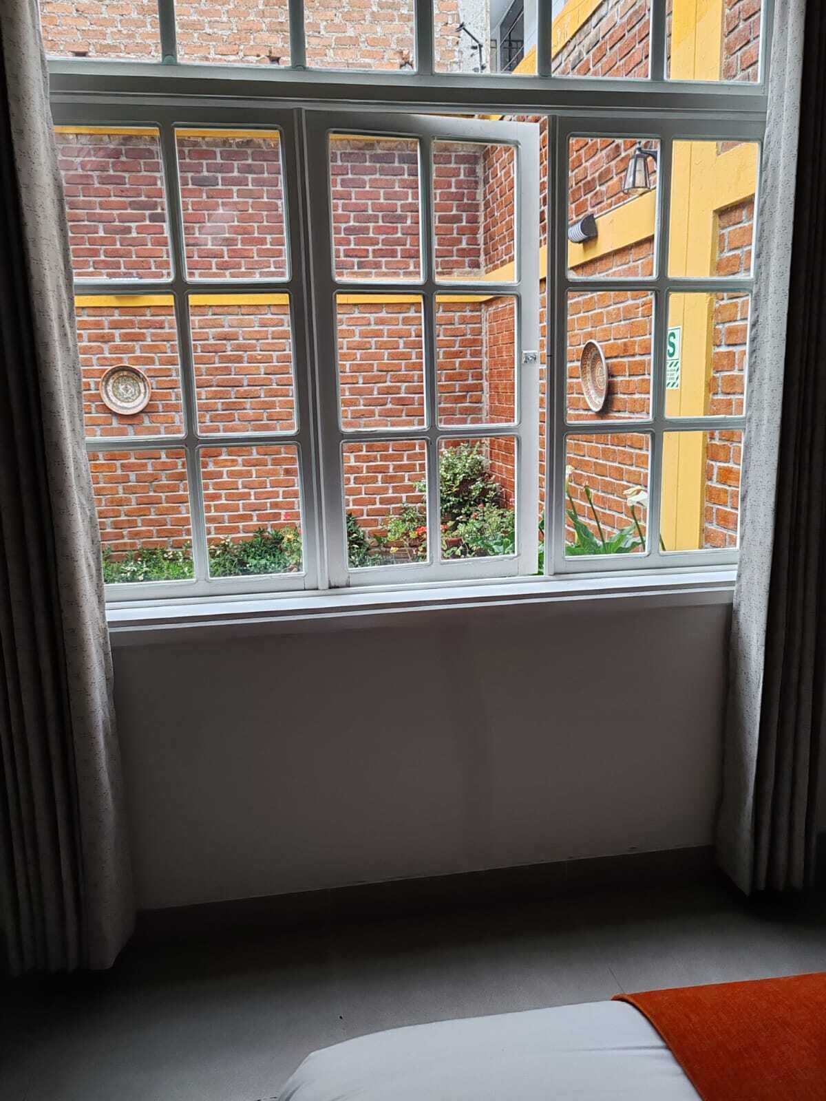 Une pièce dotée d’une grande fenêtre, d’un lit et donnant sur un immeuble en briques avec une porte jaune.