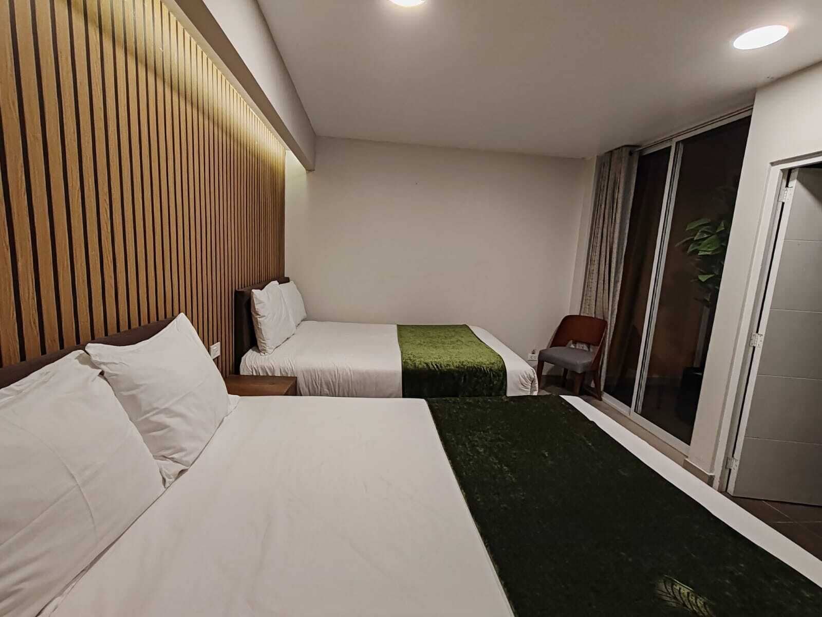 Habitación de hotel con dos camas, cabecera de madera, alfombra verde decorativa, una silla y puerta de vidrio corredera.