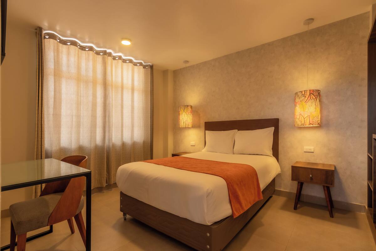 A hotel room with a bed, a bedside table, a chair, a glass table, and curtains.