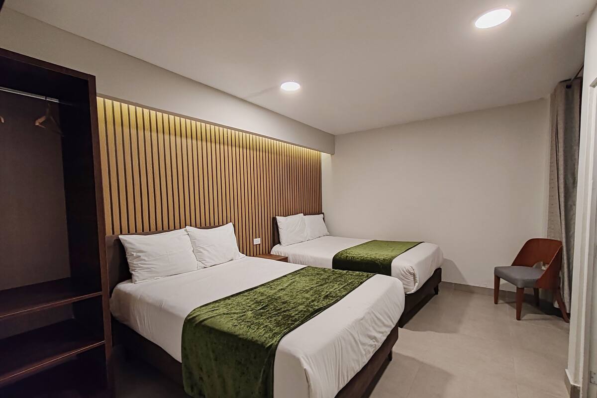 A hotel room with two beds, a wooden headboard, a green throw blanket, and a chair.