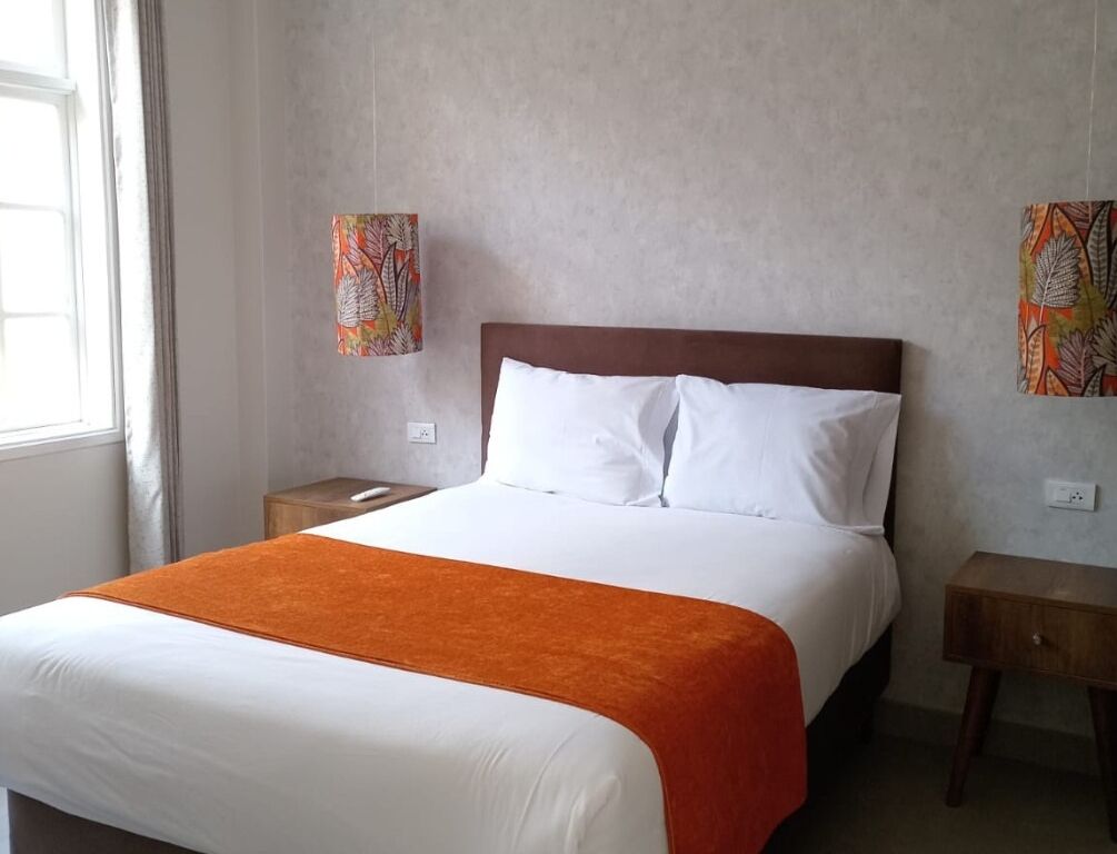 A hotel room with a bed, a nightstand, a bedside table, and a window.