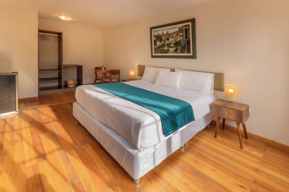 A hotel room with a large bed, wooden flooring, a wardrobe, a desk, and a painting on the wall.